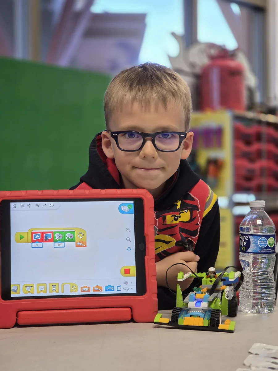 A child showing a tablet program beside a small LEGO® robot at a building table.