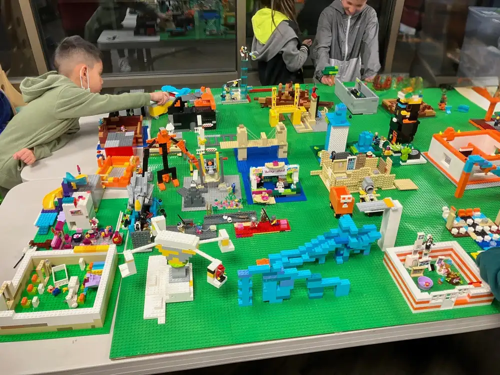 Children assembling a shared LEGO® town layout on a green building table.